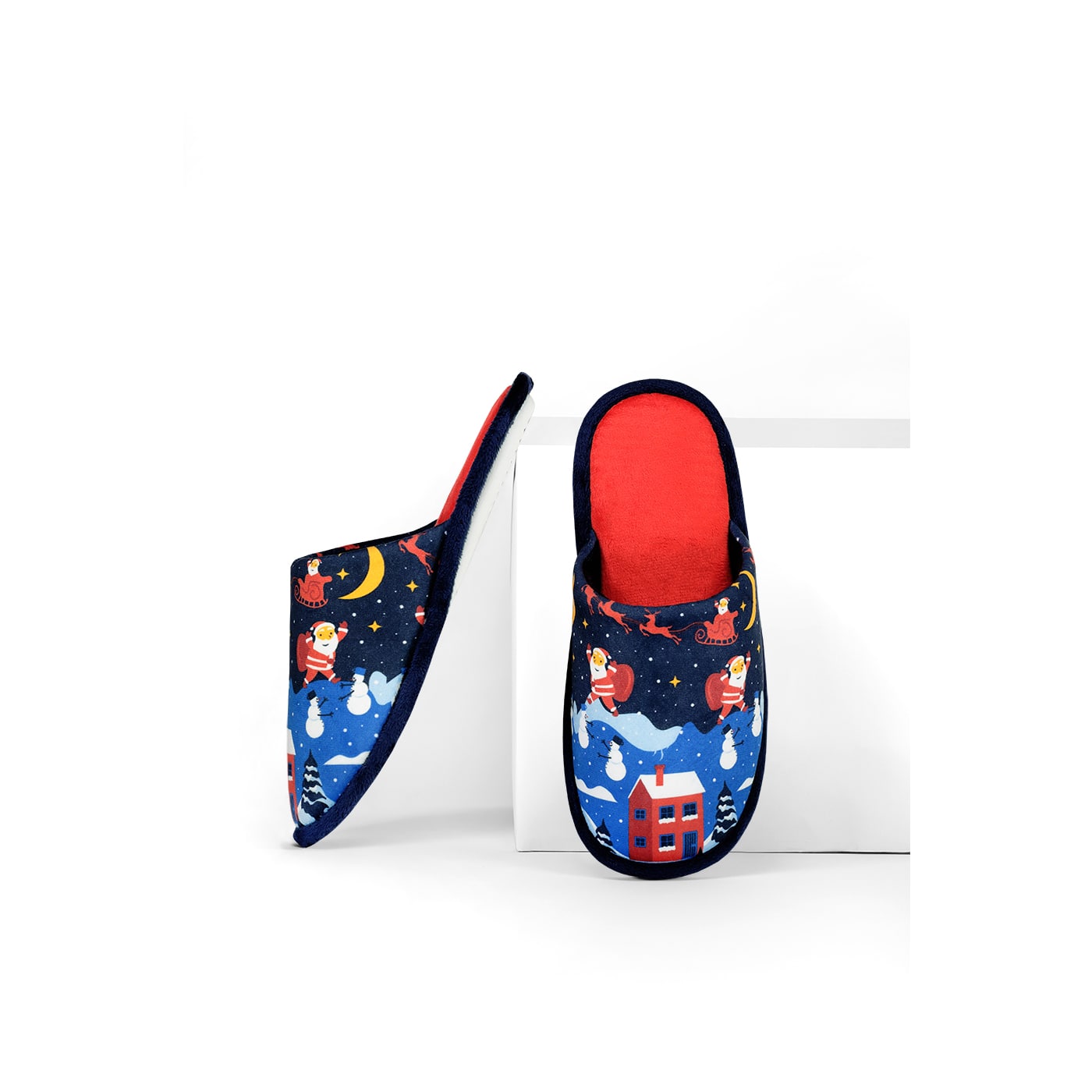 Papuci de casă cu model de Crăciun, imprimeu cu Moș Crăciun și peisaj de iarnă, pe fundal albastru, cu interior roșu, fotografiați pe fundal alb.