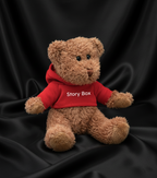 Ursuleț de pluș bej îmbrăcat cu hanorac roșu StoryBox, fotografiat pe fundal negru satinat, accesoriu din seturile cadou StoryBox.